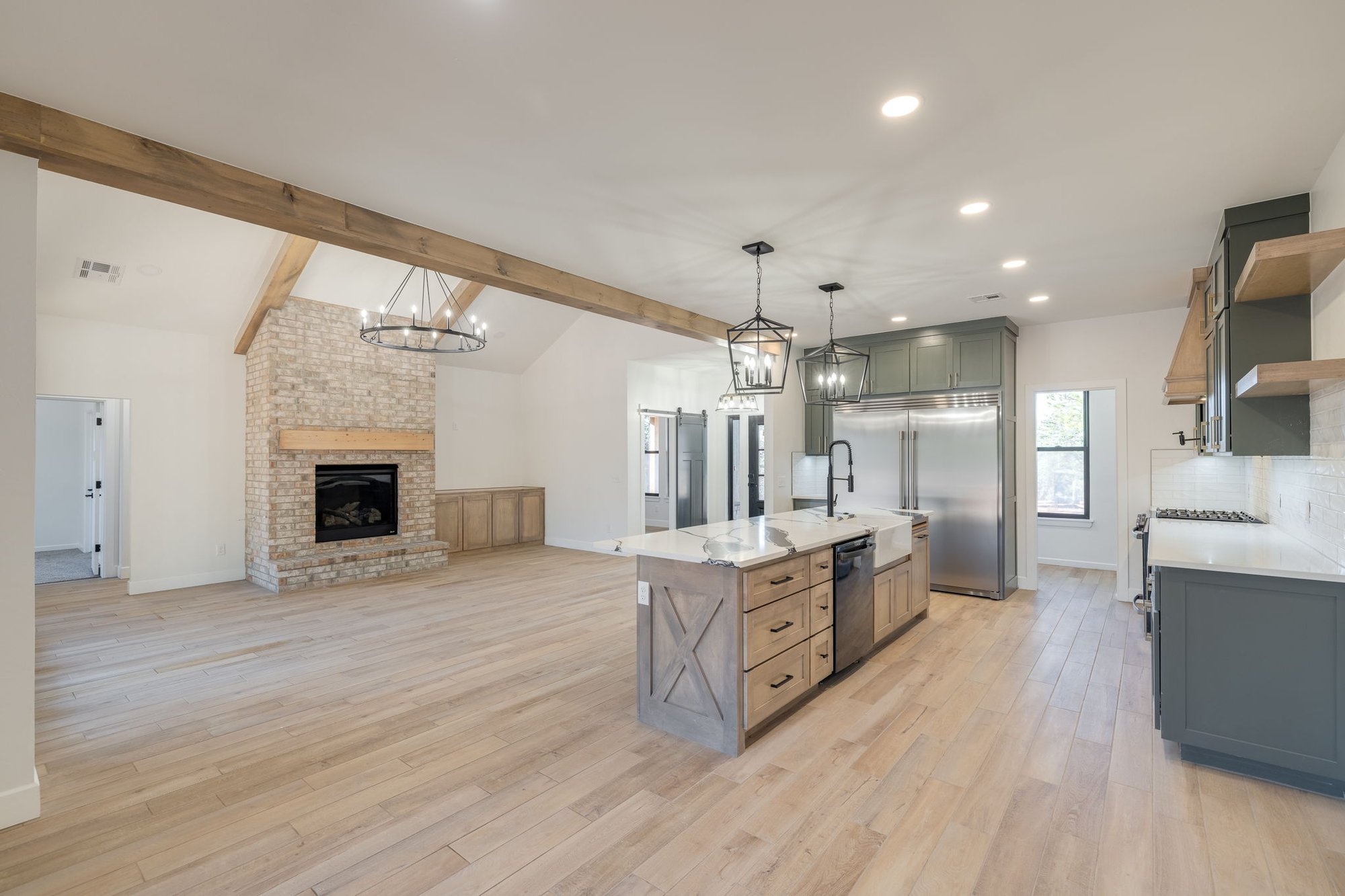 Modern farmhouse-style space featuring exposed wooden beams, a stone fireplace, and a large kitchen island with pendant lighting.