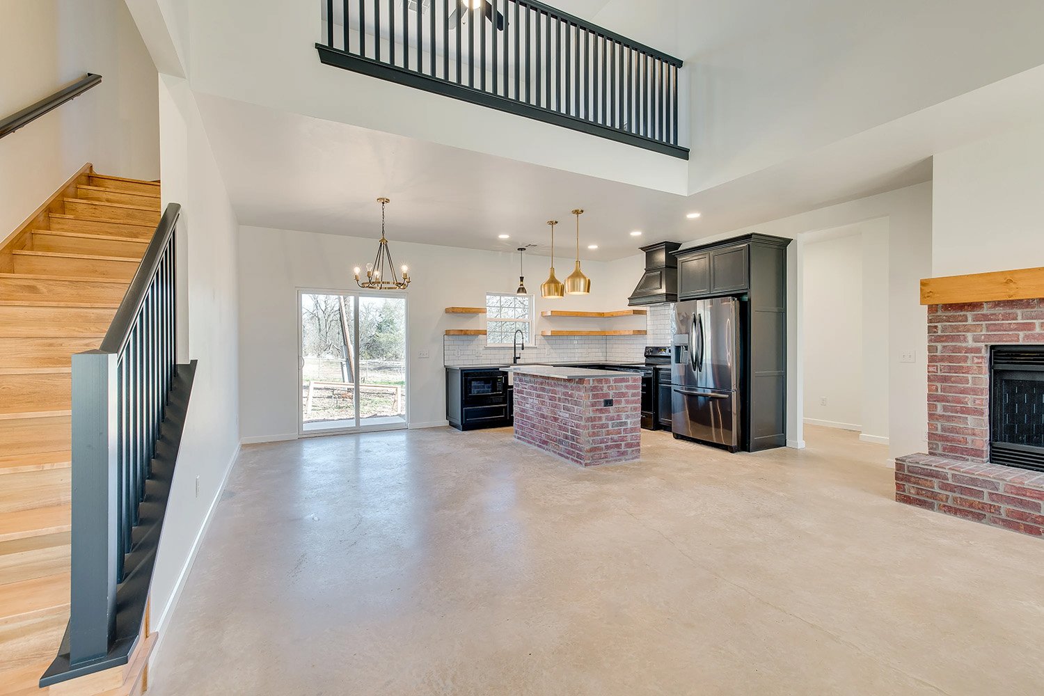 Open-concept kitchen and living space in Edmond, Oklahoma with a brick island, black cabinetry, gold pendant lighting, exposed wood shelving, and a cozy fireplace accented with red brick.