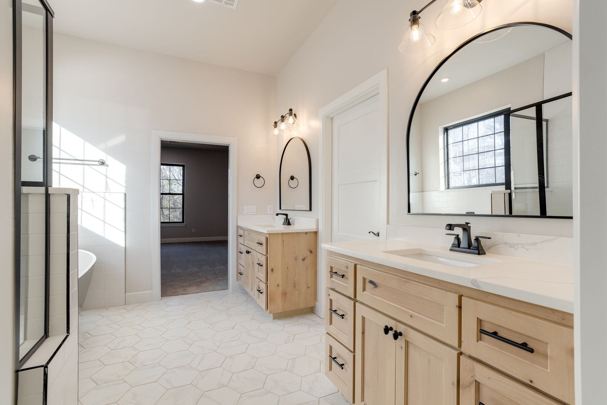 Light wood cabinets, black fixtures, arched mirrors, and hexagon tile flooring.