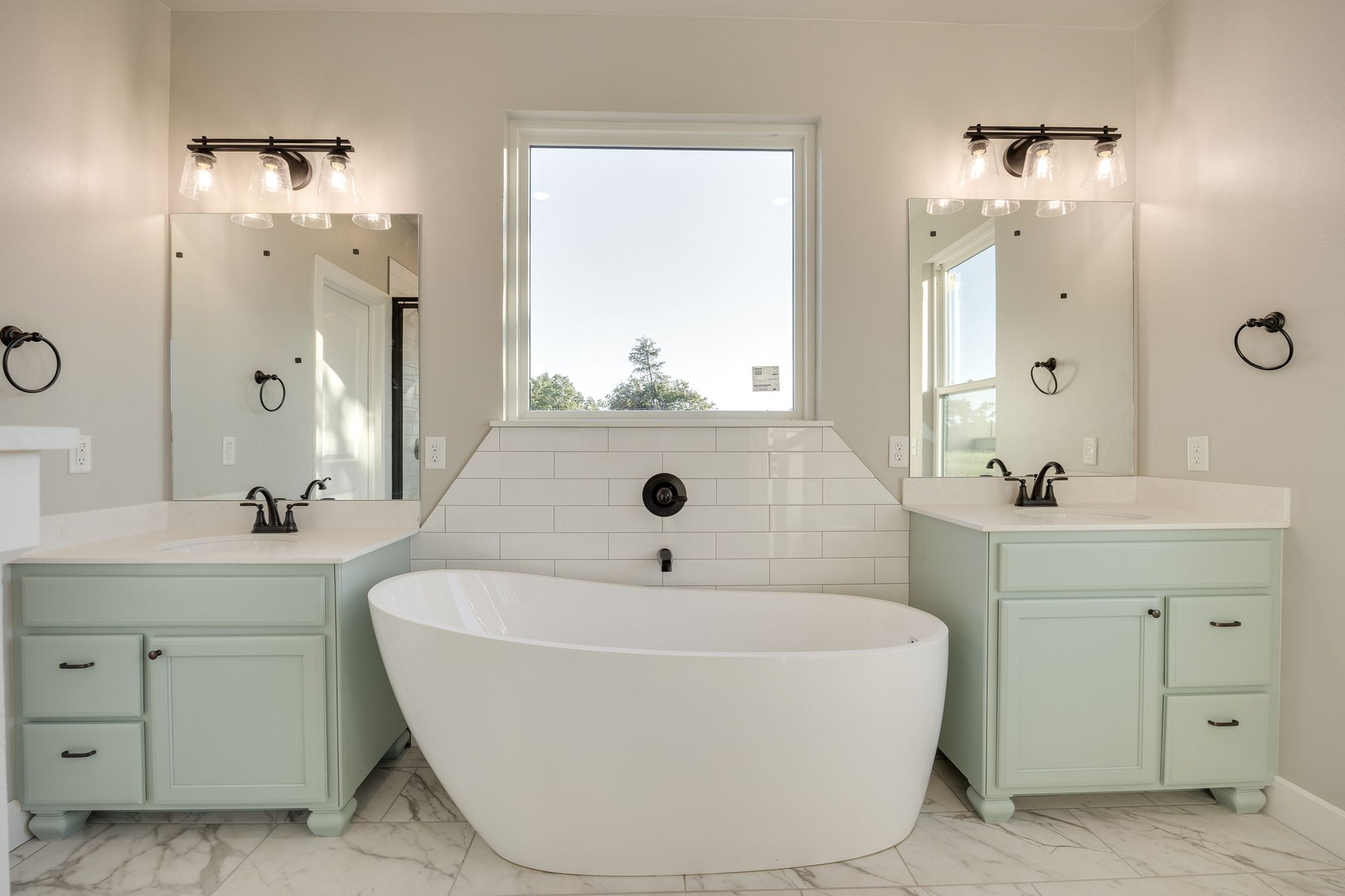 Modern bathroom in Guthrie, Oklahoma, featuring a freestanding white bathtub, dual vanities with mint green cabinets, marble tile flooring, and a large window with natural light.