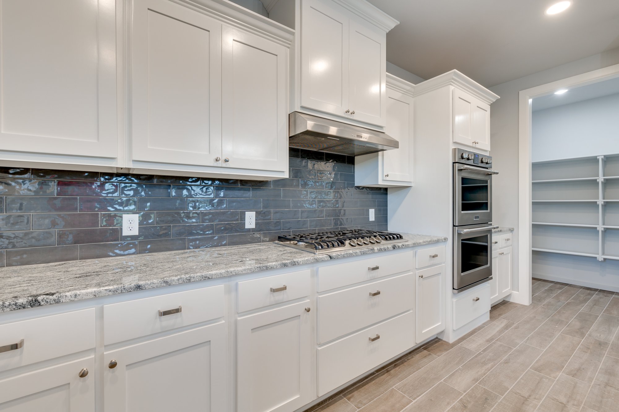 Modern kitchen in Slaughterville, Oklahoma, featuring white cabinetry, stainless steel appliances, gray tile backsplash, and granite countertops with a walk-in pantry.