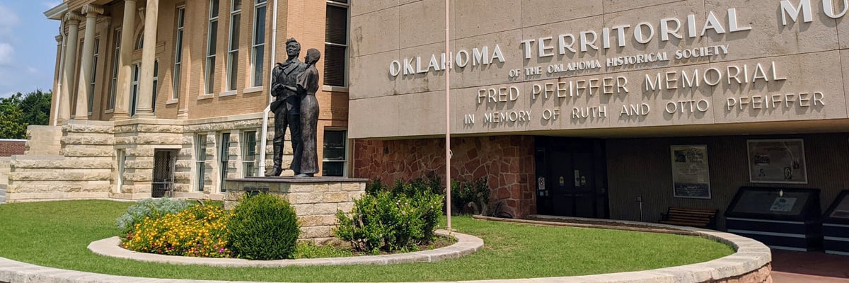 Exterior of the Oklahoma Territorial Museum in Guthrie, Oklahoma, featuring a bronze statue and landscaped flower beds.